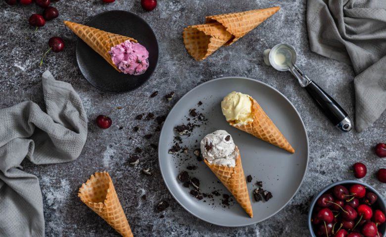 Flat lay of Chapman's Ice Cream in sugar cones along with ice cream spoon and black cherries.