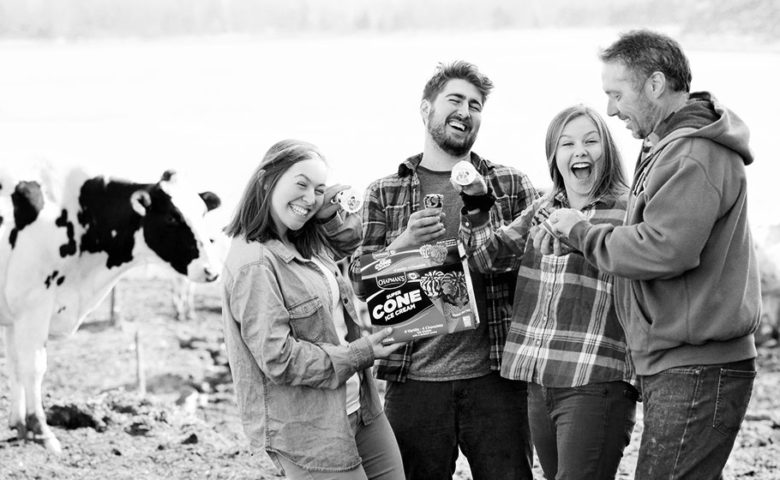 A father with his three children, who are all smiling and laughing and holding Super Cones, who are dairy farmers and standing in a barnyard with dairy cows standing on either side.