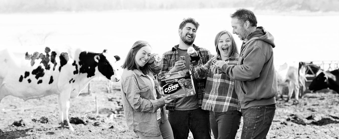 A father with his three children, who are all smiling and laughing and holding Super Cones, who are dairy farmers and standing in a barnyard with dairy cows standing on either side.