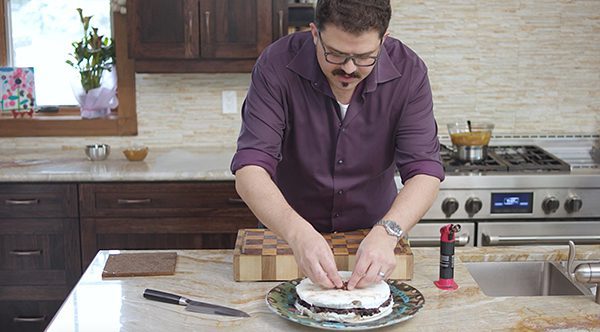 Man in purple shirt topping a Chapman's Salty Caramel cake with chocolate