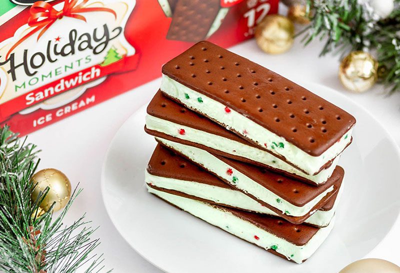 A stack of five Chapman's Peppermint Ice Cream Sandwiches on a white plate, sitting on a white table top with the product packaging blurry in the background and pine tree springs and golden Christmas balls beside the plate as decoration