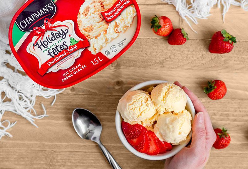 Chapman's creamy Shortbread Ice Cream in a white bowl placed on a wooden table top with fresh strawberries scattered around and the product packaging off to the side with a spoon beside the bowl.