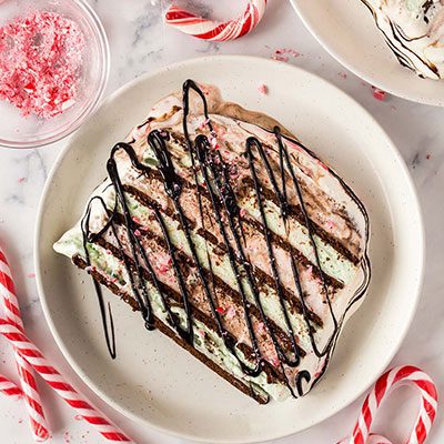 A slice of Holiday Ice Cream Cake laying on a white plate with chocolate syrup drizzled on top and candy canes and crushed candy canes surrounding the plate.