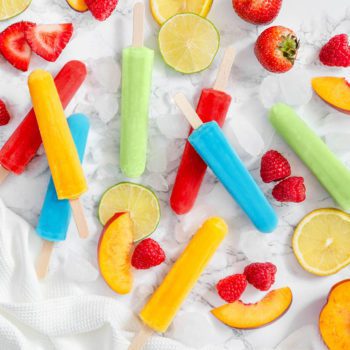 Assorted flavours of Chapman's Fruity Lolly lying on a marble table top with slices of fresh peaches, raspberries and citrus fruit