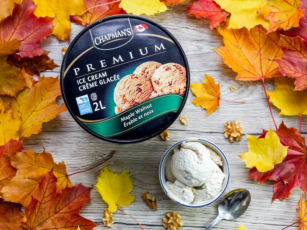 Viewed from above, a 2L tub of Chapman's Premium Maple Walnut ice cream beside a bowl filled with scoops of this flavour, on top of a light coloured wooden table with fall leaves scattered around.