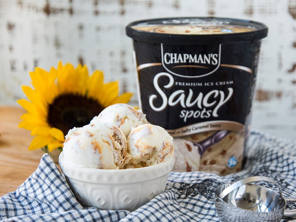 Trois grandes boules de crème glacée points saucy caramel de Chapman dans un bol blanc, à côté du contenant de produit avec un tournesol jaune vif derrière, le tout placé sur une serviette de cuisine à carreaux bleu et blanc.