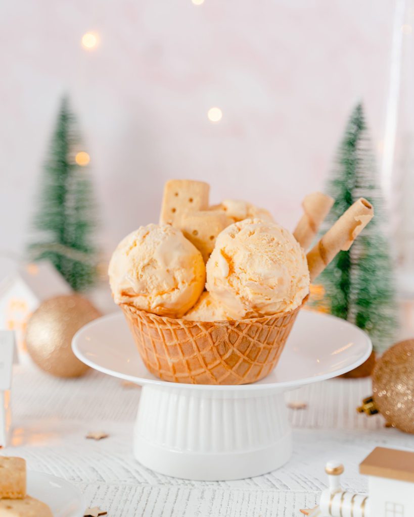 Three large scoops of Chapman's Shortbread ice cream topped with a couple shortbread cookies inside a waffle bowl in front of a winter wonderland background including golden Christmas balls, warm lights and two evergreen trees