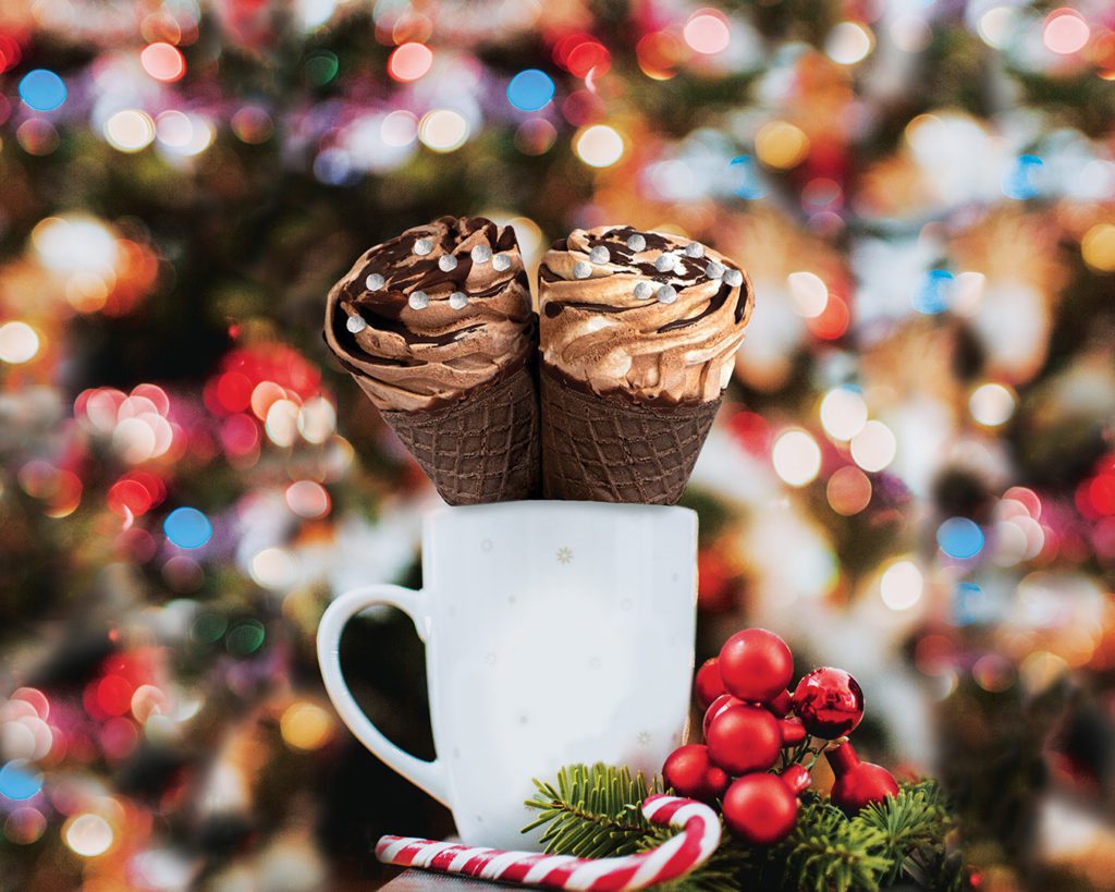 Two Chapman's Winter Cocoa ice cream cones inside a white mug with colourful Christmas lights on a tree faded in the background