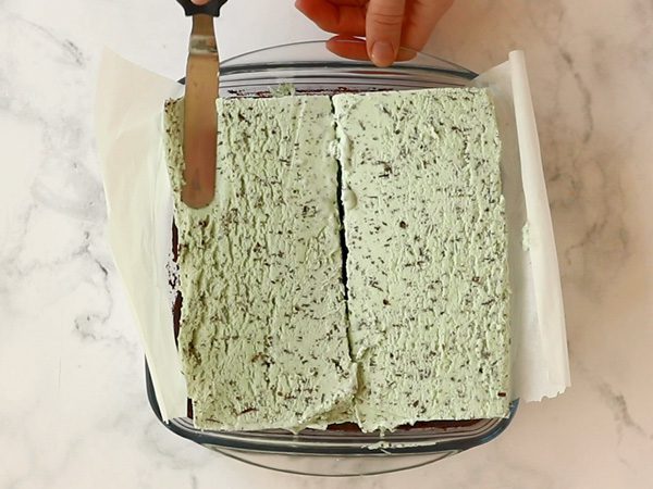 person's hand holding an offset spatula beginning to spread two large slices of Mint Chip ice cream on top of baked brownies in a square baking dish