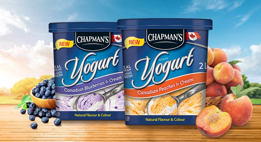 On a wooden surface, a tub of Chapman's Canadian Blueberries and Cream Frozen Yogurt with fresh blueberries to the left beside a tub of Canadian Peaches and Cream Frozen Yogurt with fresh peaches to the right.