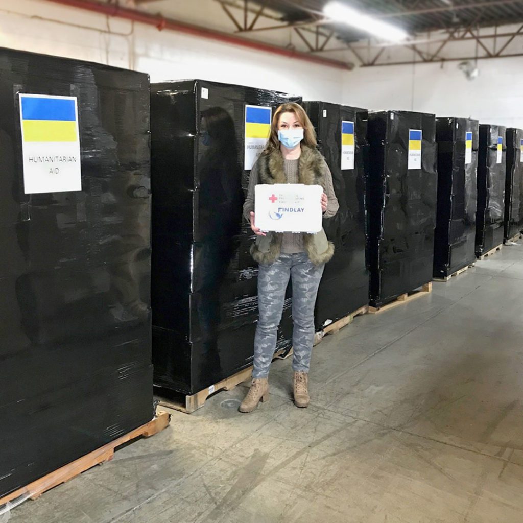 Lesya Chapman standing in front of 8 pallets full of first aid kits ready to be shipped to Ukraine