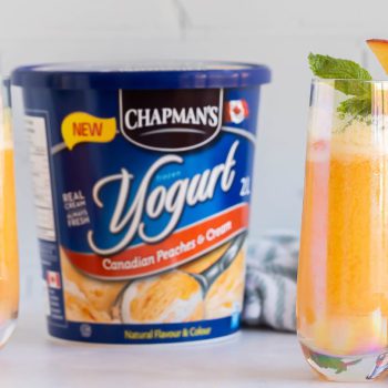 Deux grands verres remplis de mocktail bellini a la pêche à côté d’une contenant de 2 L de Yogourt Glacé pêche et crème Canadien Chapman’s sur un comptoir en marbre