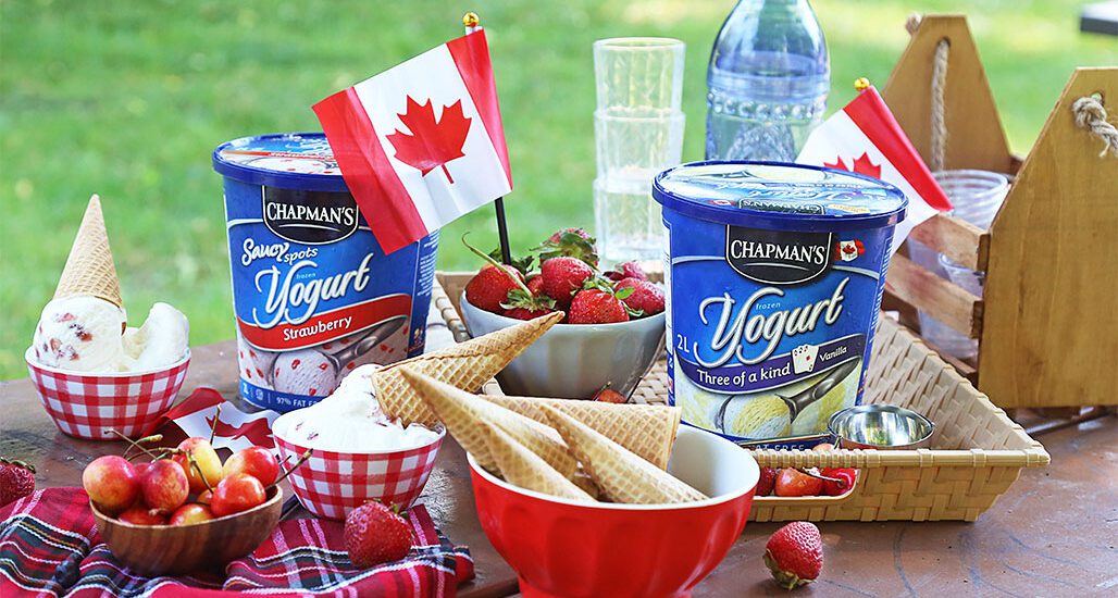 Outside with green grass in the background is a Canada Day themed spread on a wooden table. On the table are two tubs of Chapman's Frozen Yogurt, red bowls with fresh strawberries and waffle cones, and red and white checkered bowls with scoops of frozen yogurt.