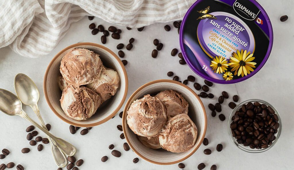 On a white counter top are two bowls of Chapman's No Sugar Added and Lactose Free Coffee Chocolate ice cream. Coffee beans are scattered around. The ice cream tub sits beside the bowls.