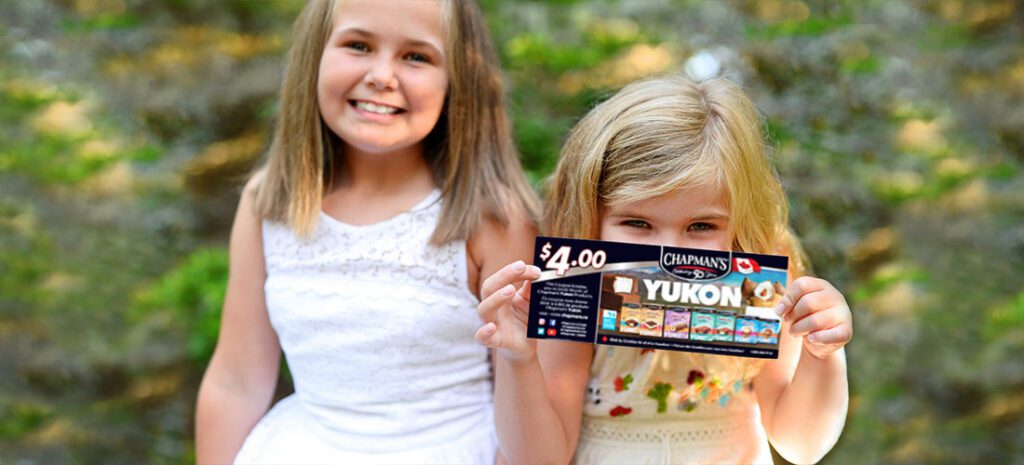 Une fille aux yeux souriants assise à l’extérieur sur du l’herbe verte tenant un coupon de 4 $ de Chapman dans les deux mains tout en couvrant partiellement son visage.