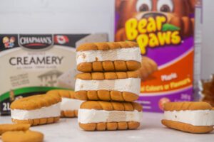 Six Maple Crunch Ice cream sandwiches on a grey table with three stacked on each other in the back ground there is packaging from Chapman's and Bear Paws.