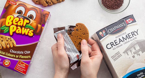 On a light grey countertop lays a box of Bear Paws Chocolate Chip cookies and a carton of Chapman's Markdale Creamery ice cream. In the center of the photo are two hands opening up the wrapper of a Bear Paws cookie and taking a cookie out.