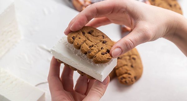 View of someone's hand placing a slice of Chapman's Vanilla ice cream between two Bear Paws Chocolate Chip cookies.