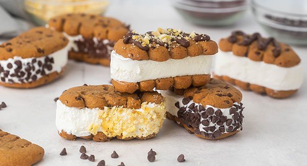 A stack of three Chocolate Chip Ice Cream Sandwiches on a light grey countertop. The sandwiches have been garnished with yummy toppings like mini chocolate chips and crushed banana chips. Behind the stack of sandwiches are three more sandwiches out of focus.