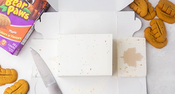 A carton of Chapman's Maple Crunch ice cream fully opened up on a white marble countertop. Around the carton's flaps are a few Bear Paws cookies and the packaging.