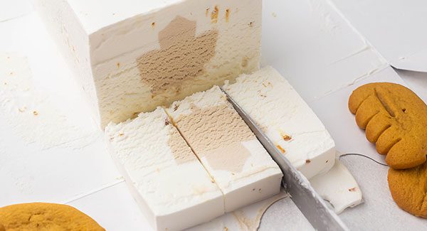 The end of a brick of Chapman's Maple Crunch ice cream the has been sliced to reveal a maple leaf in the center. A knife is cutting a slice of ice cream that is laying on the edge of the carton into thirds.