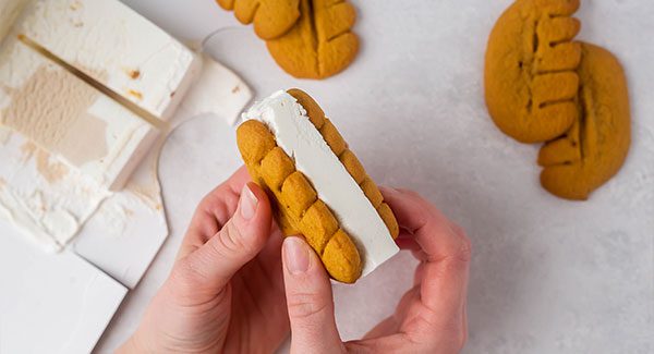 On a white marble countertop is an open carton of Maple Crunch ice cream with a slice of ice cream cut into thirds and four Bear Paws cookies. Two hands are seen pressing together a slice of ice cream in between two Bear Paws cookies.