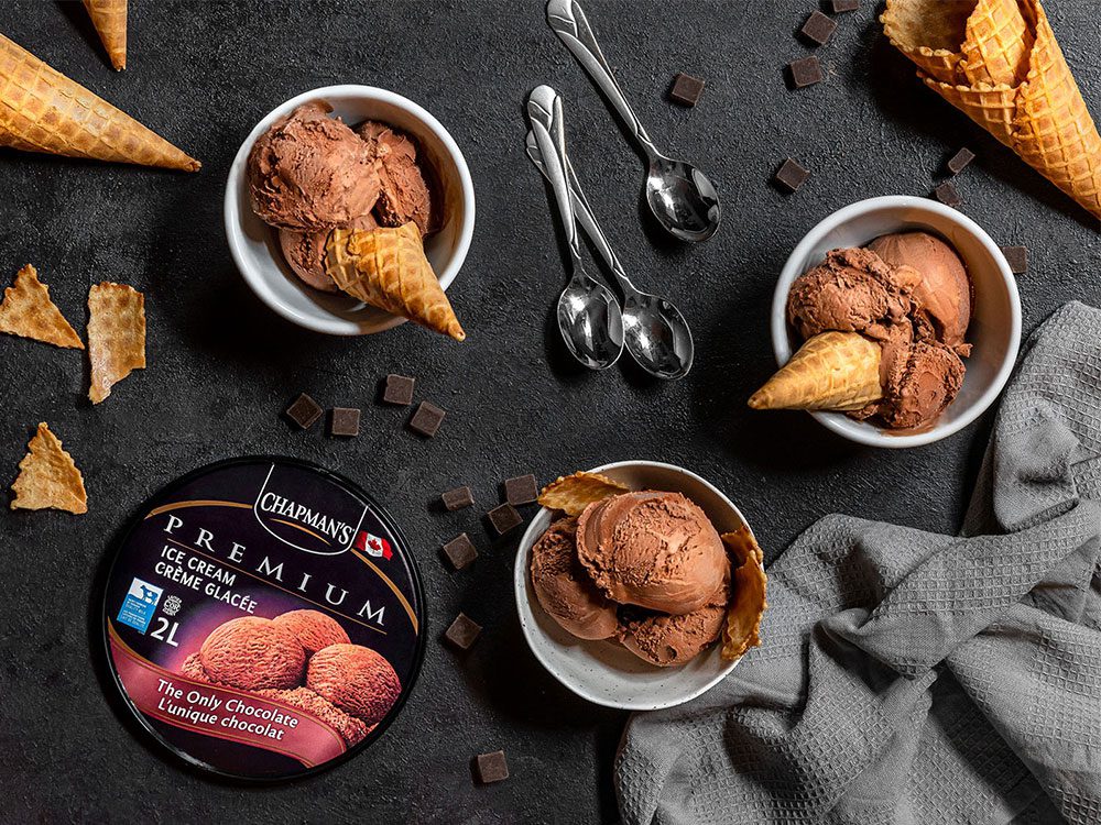 Three bowls of Chapman's The only Chocolate with sugar cones as garnish on a black table top covered in sugar cones small pieces of chocolate with a grey sweater and a tub of The only chocolate Ice Cream