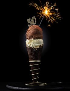 A chocolate Ice Cream cone with a lit sparkler and a 50 on top of the ice cream in a ice cream holder with a black background