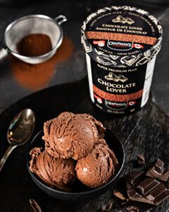 A chocolate bowl of ice cream in a black bowl with three scoops on a dark tabple top with a spoon little pieces of chocolate, coco in a sifter and a tub of Chocolate lover Ice Cream