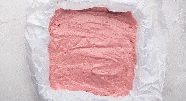 The pink batter has been put into a baking pan lined with wax paper