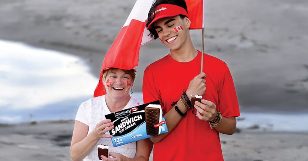A smiling grandma and her grandson standing on a beach in Eastern Canada are holding a Canadian flag and a box of Chapman's Vanilla Ice Cream Sandwiches.