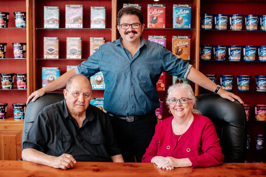 With Ashley Chapman standing behind them, David and Penny Chapman are sitting at a table in Chapman's head office boardroom. An array of Chapman's products are on display behind them.