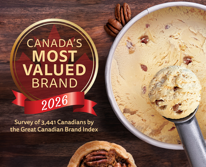 An overhead view of an open tub of Chapman's Pecan Butter Tart ice cream on a dark wood table top. A large scoop of ice cream is held over the tub. A pecan butter tart sits on the wooden surface, along with a couple pecans. The Canada's Most Valued Brand 2026 award badge graphic overlays the dark wooden surface to the left of the tub.