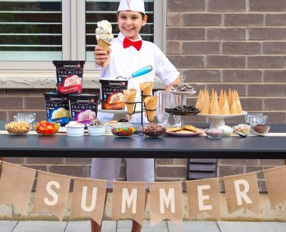 A Child is behind a self made ice cream counter. The counter is full of Ice Cream, Ice Cream cones, and various ice cream toppings. There is a banner below the table that says summer